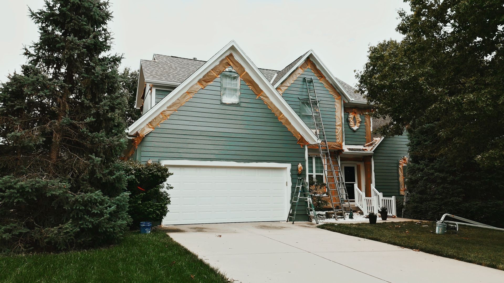 Two-story house being painted with ladders and equipment in the front, surrounded by trees.